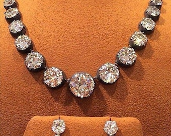 May include: A dazzling diamond necklace and matching earrings. The necklace features large, round-cut diamonds set in a dark metal. The earrings have a similar design, with a smaller diamond above a larger one. The jewelry is displayed on a brown surface.
