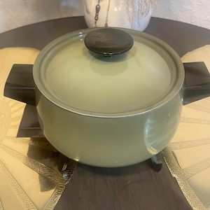 May include: A vintage, pale green cooking pot with a matching lid and a dark brown handle. The pot has black handles on either side. The pot is sitting on a dark brown surface.