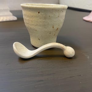 May include: A small, off-white ceramic cup with a textured surface and a matching spoon. The cup has a flared shape, and the spoon has a curved handle with a rounded end. The items are likely for serving a small portion of food.