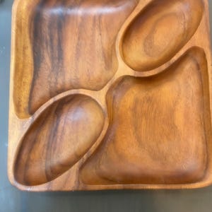 Puede incluir: Una bandeja de madera cuadrada con cuatro compartimentos separados. La bandeja está hecha de madera de color marrón claro con un patrón de vetas natural. Los compartimentos tienen varias formas orgánicas, perfectos para servir aperitivos.