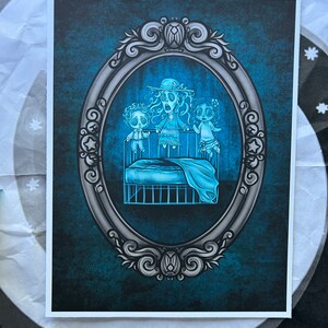 May include: A vintage-style oval frame with a blue and gray background. Inside the frame, three ghostly figures are depicted standing in a crib. The figures are translucent and have large, wide eyes. The crib is made of metal and has a blue blanket.