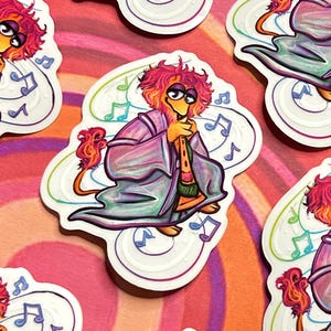May include: A colorful sticker featuring a cartoon character with red hair, a purple robe, and a tail playing a flute. The character is surrounded by musical notes.