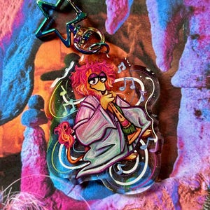 May include: A pink and purple lion character with a blue and green star charm keychain. The lion is wearing a purple robe and has a musical note design around it.