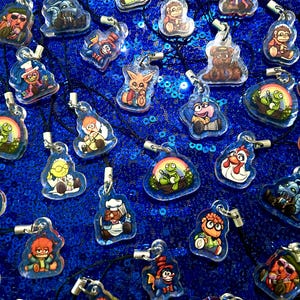 May include: A collection of clear acrylic character pendants on black cords, featuring various cartoon characters. The pendants are displayed on a shimmering blue sequined surface. Each pendant has a silver-colored clasp.