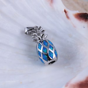 Op de afbeelding: Een zilveren ananasbedel met blauwe opaal-inleg. De bedel heeft een lus aan de bovenkant om aan een ketting of armband te bevestigen.