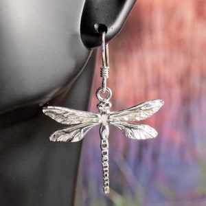 May include: Silver dragonfly earrings with detailed wings and a delicate hook. The earrings are suspended from a black surface, with a blurred background of purple and pink hues. The dragonfly design is a classic and elegant jewelry choice.