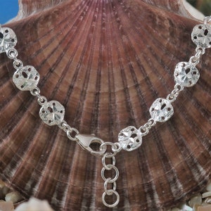 Puede incluir: Un brazalete de plata con una cadena y siete dijes de dólar de arena. El brazalete está sobre un fondo de concha de mar marrón.