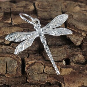 Könnte beinhalten: Ein silberner Libellen-Anhänger mit detaillierten Flügeln und einem strukturierten Körper. Der Anhänger ist so konzipiert, dass er als Anhänger oder Perle für die Schmuckherstellung verwendet werden kann.