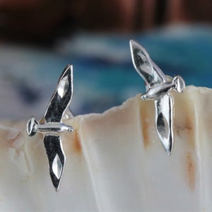 May include: A pair of silver-colored stud earrings shaped like seagulls in flight. The earrings have detailed wings and bodies, with a polished finish. The earrings are displayed on a white, textured surface, with a blurred blue and white background.