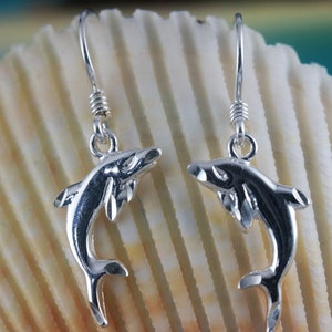 Puede incluir: Un par de pendientes de plata con forma de delfín con un diseño colgante. Los delfines miran hacia adelante con las colas enroscadas.