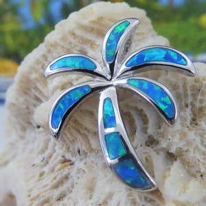 May include: A silver palm tree pendant with blue opal inlay. The pendant is on a white coral-like surface.