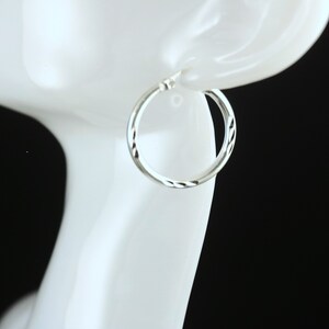 May include: Silver hoop earrings with a textured design. The earrings are shown on a white mannequin head.
