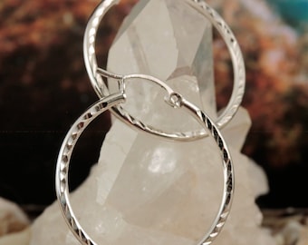 May include: A pair of silver hoop earrings with a textured diamond pattern. The earrings are displayed on a white crystal background.