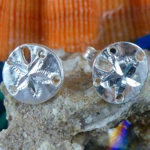 May include: A pair of silver sand dollar earrings. The earrings are round and have a detailed design of a sand dollar with a five-pointed star in the center.