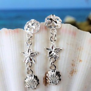 May include: Silver dangle earrings featuring a sand dollar, starfish, and seashell charm. The earrings are displayed on a seashell.