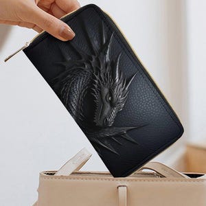 May include: Black wallet with a detailed dragon design. The wallet is rectangular with a gold zipper and is being placed into a beige handbag. The dragon is embossed on the front of the wallet.