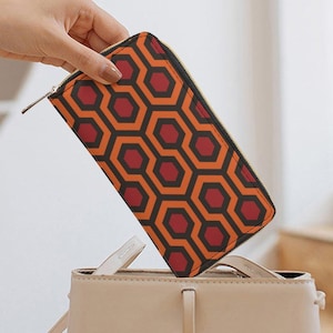 May include: A rectangular wallet with a zipper closure and a repeating geometric pattern of orange and red hexagons on a black background. The wallet is being placed into a beige handbag.