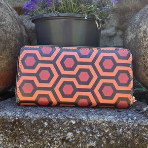 May include: A rectangular zip-around wallet featuring a repeating geometric pattern of interlocking hexagons in shades of orange, red, and black. The wallet has a structured shape with a smooth surface and a gold-toned zipper pull visible on the side.