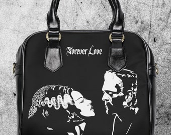 May include: Black handbag featuring a white graphic of the Bride of Frankenstein and Frankenstein, with the words "Forever Love" above the image. The bag has a top handle and gold-colored hardware.