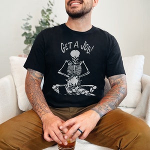 Op de afbeelding: Een zwart T-shirt met een witte skeletprint en de tekst "GET A JOB!" gedrukt op de voorkant. Het skelet zit in een meditatieve houding met een schedel op zijn schoot.
