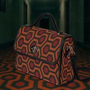 May include: A satchel handbag featuring a repeating geometric pattern in orange and burgundy. It has a black handle and strap, with a silver clasp. The bag is placed on a patterned carpet.