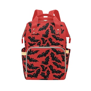 Peut inclure: Un sac à dos rouge orné d'un motif répétitif de chauves-souris noires. Le sac comprend une poignée supérieure, deux bretelles et une poche frontale zippée. Le motif est uniforme sur le corps principal et la poche avec des détails dorés.
