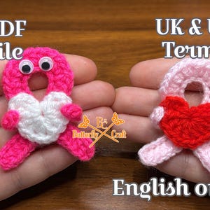 May include: Two handmade crochet heart-shaped ornaments. One is pink with a white heart and googly eyes, the other is pink with a red heart. The text "PDF file" and "UK & US Terms" are visible.