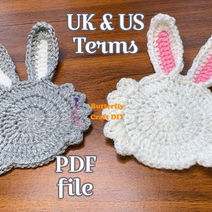 Peut inclure: Deux dessous-de-verre en crochet en forme de lapin, l'un gris et l'autre blanc, avec des oreilles de lapin roses. Les dessous-de-verre sont sur une surface en bois. Le texte "UK & US Terms" et "PDF file" est visible sur l'image.