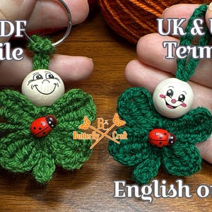 May include: Two handmade keychains featuring a green crocheted four-leaf clover design with a wooden smiling face and a red ladybug. The keychains are attached to a silver keyring. The text "PDF file" and "UK & US Terms" are visible.