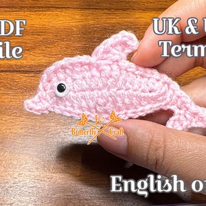 May include: A light pink crocheted dolphin applique featuring a single black and white plastic googly eye. The image includes text that reads PDF file, UK & US Terms, Butterfly Craft, and English only.