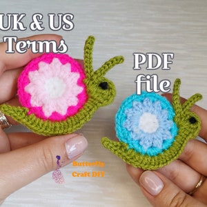 May include: Two crocheted snail amigurumi toys. One snail is green with a pink and white flower, the other is green with a blue and white flower. The text "UK & US Terms" and "PDF file" is visible in the image.