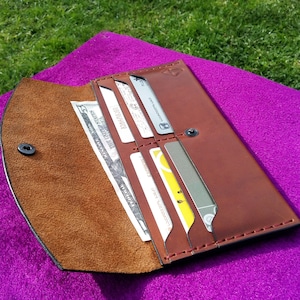 Puede incluir: Una cartera de cuero marrón con cierre de solapa y múltiples ranuras para tarjetas. La cartera está abierta y tiene un portafolios y varias tarjetas en su interior.