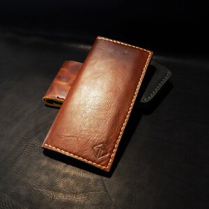 Puede incluir: Una cartera de cuero marrón con un logotipo de diamante y un borde cosido. La cartera está cerrada y se encuentra sobre una superficie marrón oscura.