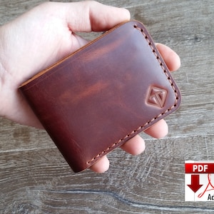 May include: A brown leather wallet with a stitched border and a geometric logo. The wallet is held in a hand, showcasing its rich color and texture. The wallet appears to be approximately 4 inches wide and 3 inches tall.
