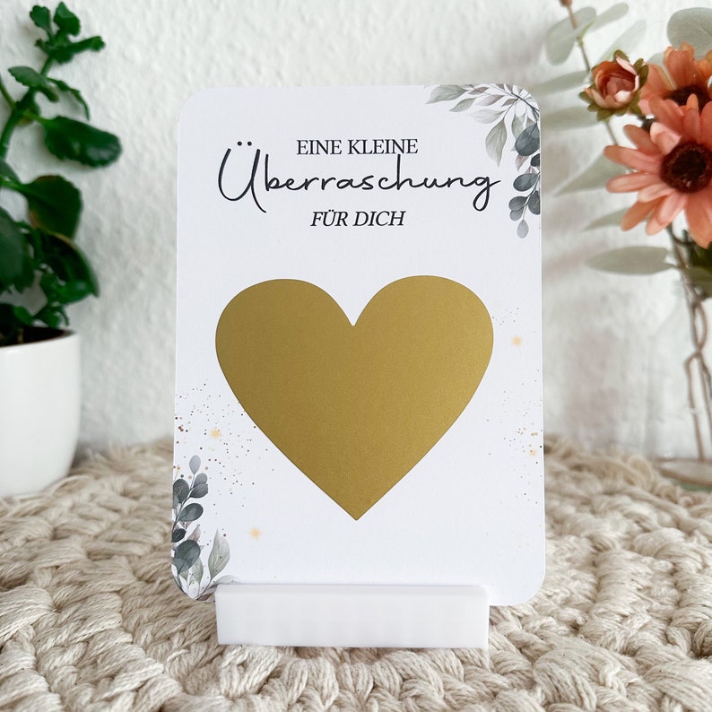 Op de afbeelding: Een witte kaart met een gouden hart en de tekst "Eine kleine &Uuml;berraschung f&uuml;r dich" wat "Een kleine verrassing voor jou" betekent.