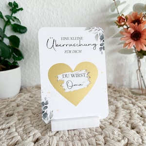Op de afbeelding: Een witte kaart met een gouden hart en de tekst "Eine kleine &Uuml;berraschung f&uuml;r dich" en "Du wirst Oma" erop. De kaart staat op een witte standaard en heeft een bloemmotief.