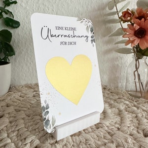 Op de afbeelding: Een witte kaart met een gouden hart en de tekst "Eine kleine &Uuml;berraschung f&uuml;r dich" in zwarte letters. De kaart is versierd met groene bladeren en gouden glitter. De kaart staat op een witte standaard.