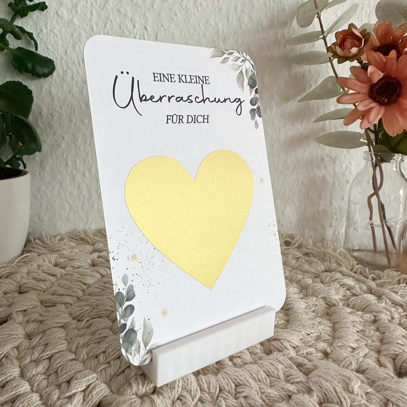 Op de afbeelding: Een witte kaart met een gouden hart en de tekst "Eine kleine &Uuml;berraschung f&uuml;r dich" in zwarte letters. De kaart is versierd met groene bladeren en gouden glitter. De kaart staat op een witte standaard.
