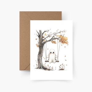 Op de afbeelding: Een witte kaart met een aquarel illustratie van een spook op een schommel onder een boom met herfstbladeren. De kaart wordt geleverd met een bruine envelop, perfect voor Halloween of herfstthema's.