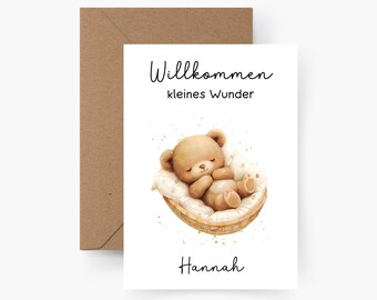 May include: A white card with the text "Willkommen kleines Wunder" and the name "Hannah" at the bottom. A brown teddy bear is sleeping in a wicker basket with a white blanket.