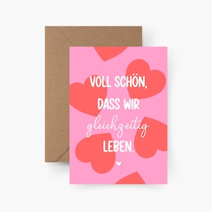 May include: A rectangular greeting card with a light pink background featuring multiple red hearts and the white text "VOLL SCHÖN, DASS WIR gleichzeitig LEBEN." A brown kraft paper envelope is positioned behind the card, partially visible on the left side.