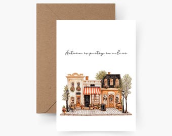 May include: Greeting card with an illustration of quaint buildings in autumn colors. The card features a brown envelope and the text "Autumn is poetry in colour". The buildings have awnings, flower boxes, and a small chalkboard sign.