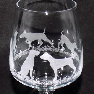 May include: A clear glass wine glass with a stem and a round base. The glass is etched with a design of four dogs running through tall grass.