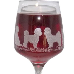 May include: A clear glass wine goblet filled with deep red liquid, featuring a frosted white etched design around the bowl that depicts a series of small dogs standing in a grassy field, with a slender stem and a circular base.