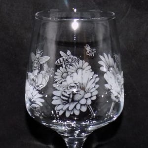 May include: A clear wine glass with etched designs of bees and flowers. The glass has a long stem and a wide bowl. The etched details add a touch of elegance to the glassware, ideal for enjoying wine. The glass is measured in centimetres.
