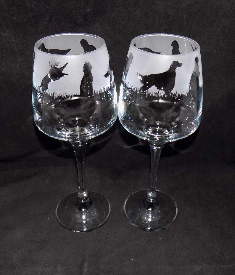 May include: Two clear glass wine glasses with etched silhouettes of dogs. The glasses have a long stem and a round bowl.