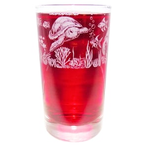 May include: A clear glass tumbler filled with a vibrant red liquid, featuring an etched design of a sea turtle swimming among coral reefs, seaweed, and various small fish. The detailed white engravings wrap around the upper portion of the glass.