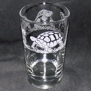 May include: A clear glass tumbler with etched turtle designs. The tumbler features a large turtle in the centre and smaller turtles around the rim. The design is white against the clear glass, creating a detailed and decorative effect.