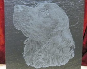 May include: Square slate coaster featuring an etched portrait of a dog's head in profile. The dog's fur is detailed with fine lines, and the image is in shades of gray against the dark slate. A decorative item for dog lovers.