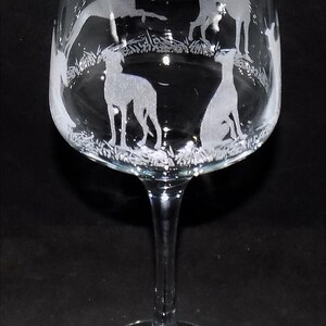 K&ouml;nnte beinhalten: Ein Weinglas aus klarem Glas mit einer ge&auml;tzten Darstellung mehrerer Windhunde in verschiedenen Posen, stehend und sitzend, umgeben von einem grasbewachsenen Landschaftsmotiv, das den unteren Teil des Glaskelchs umschlie&szlig;t.